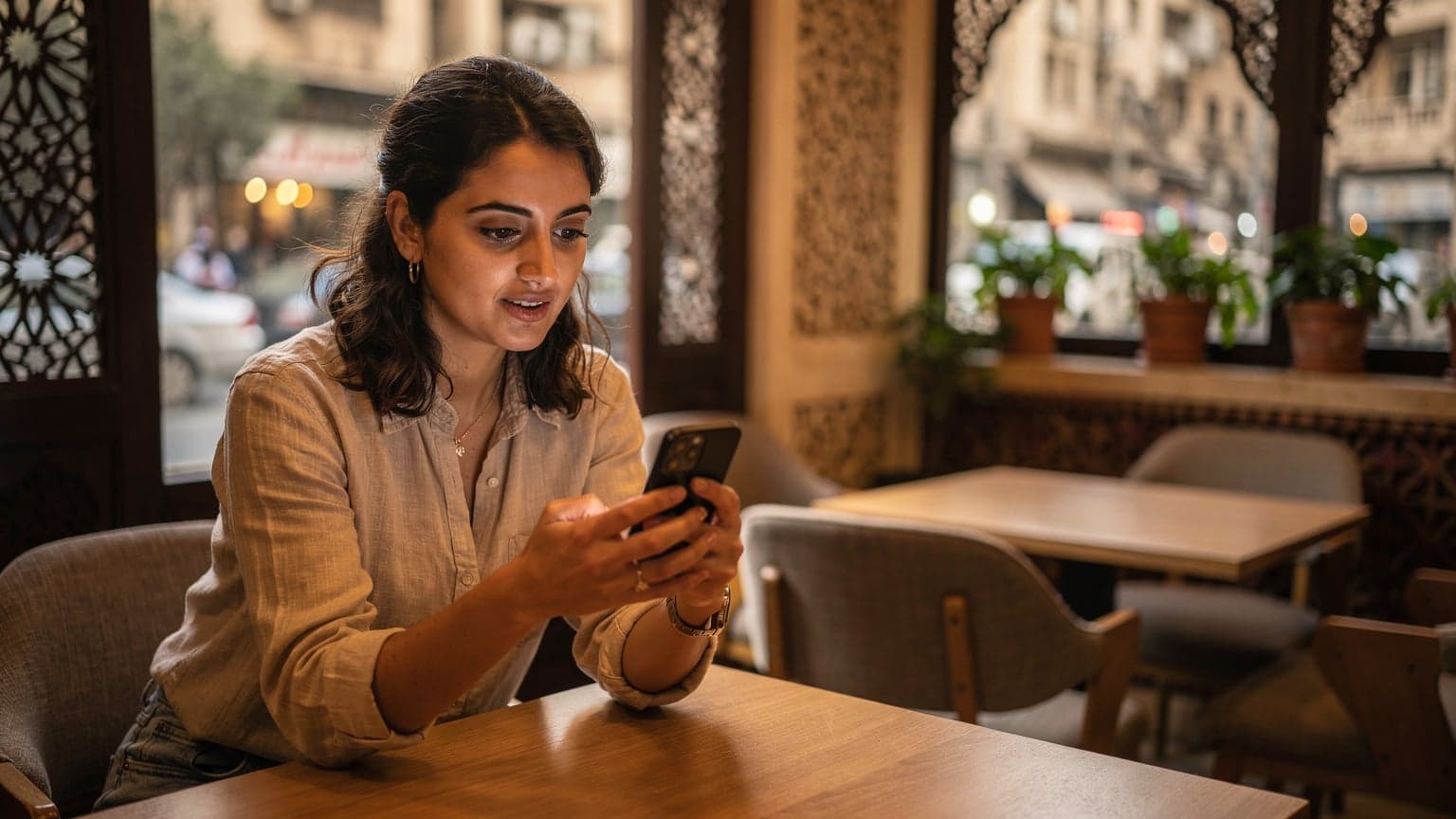 Une femme nomade numérique consultante, assise dans un café moderne du Caire avec une vue sur la ville, utilise son smartphone équipé d'une eSIM pour passer un appel vidéo, son ordinateur portable ouvert à côté d'une tasse de thé à la menthe.