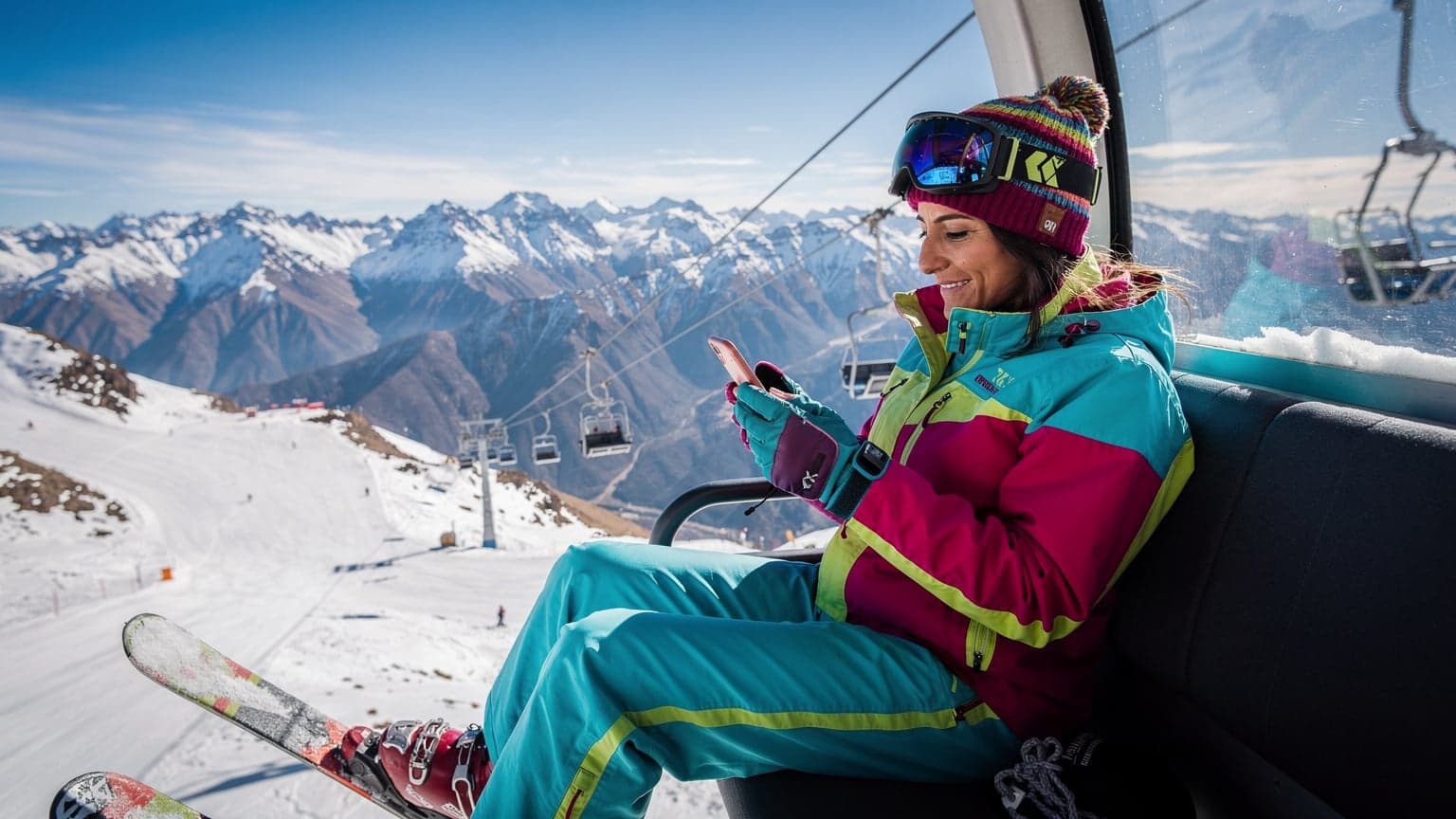 Uma mulher sorridente em roupas de esqui, sentada em um teleférico com as montanhas nevadas dos Andes ao fundo, olhando para o celular com um sorriso de satisfação.