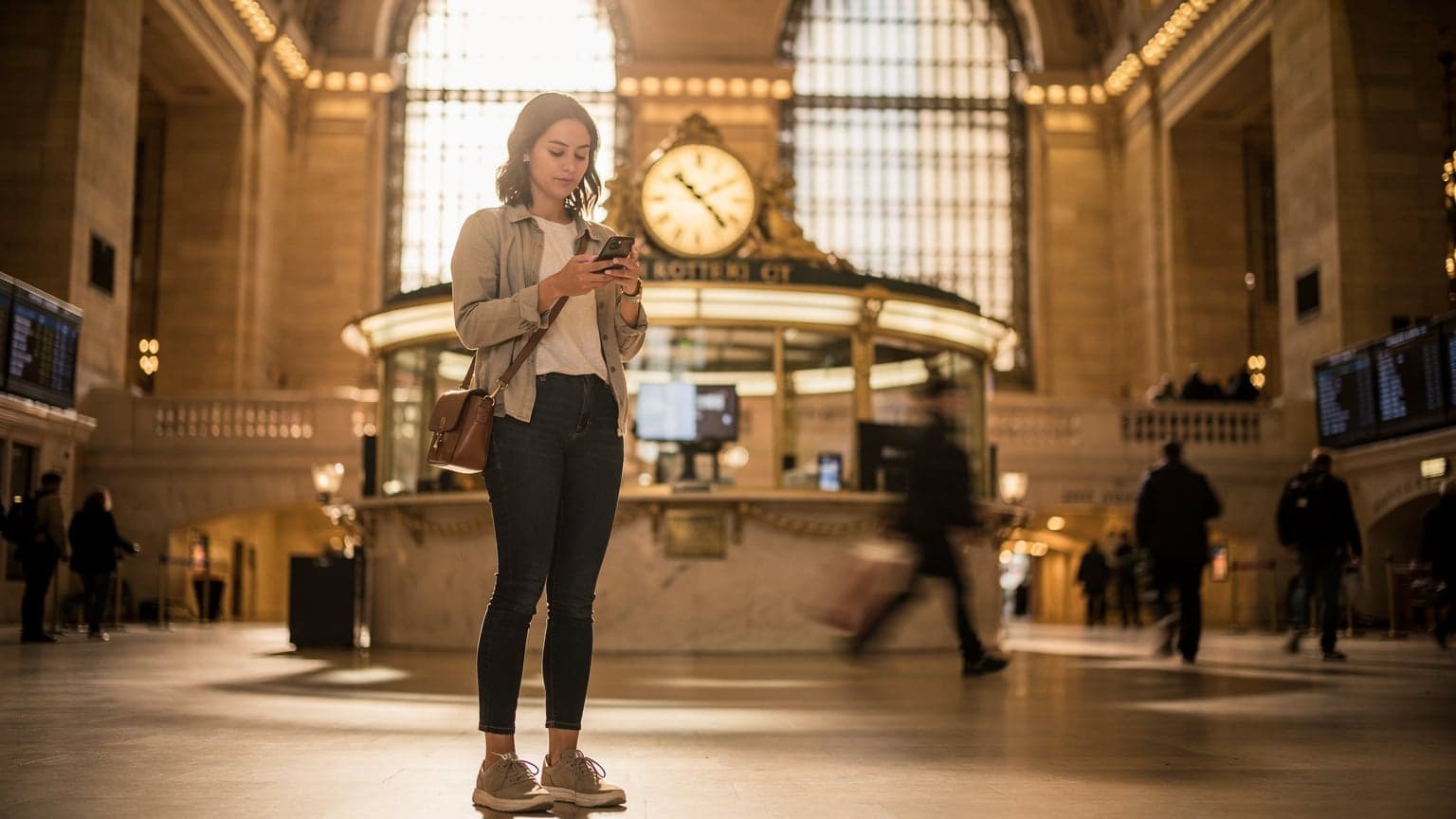 Eine junge Frau steht mit einem Smartphone in der Hand vor einer digitalen Anzeigetafel am Grand Central Terminal in New York City, während sie ihre Route überprüft.
