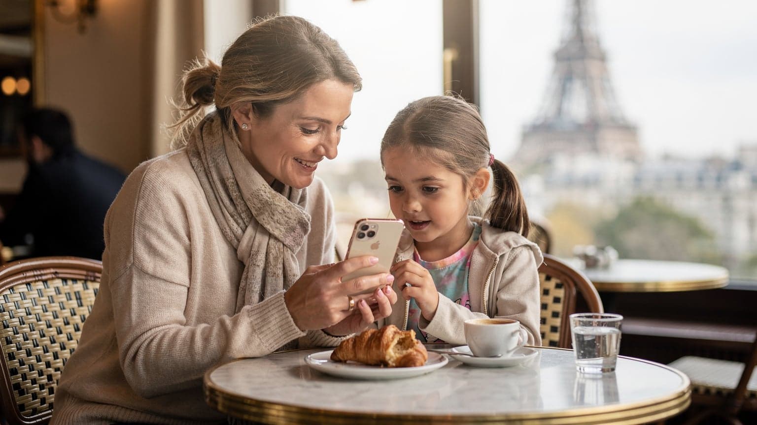 Une mère souriante et son enfant regardent un iPhone ensemble dans un café parisien lumineux, avec la Tour Eiffel visible en arrière-plan à travers la fenêtre.