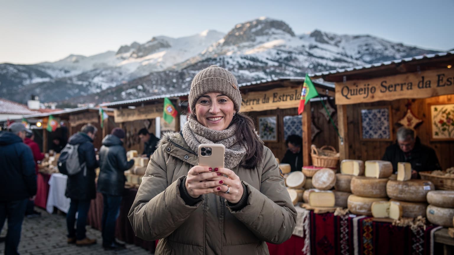 Uma mulher sorridente, vestida com um casaco quente, segura o seu smartphone e aponta para as montanhas cobertas de neve da Serra da Estrela, com uma barraca de queijo tradicional ao fundo.