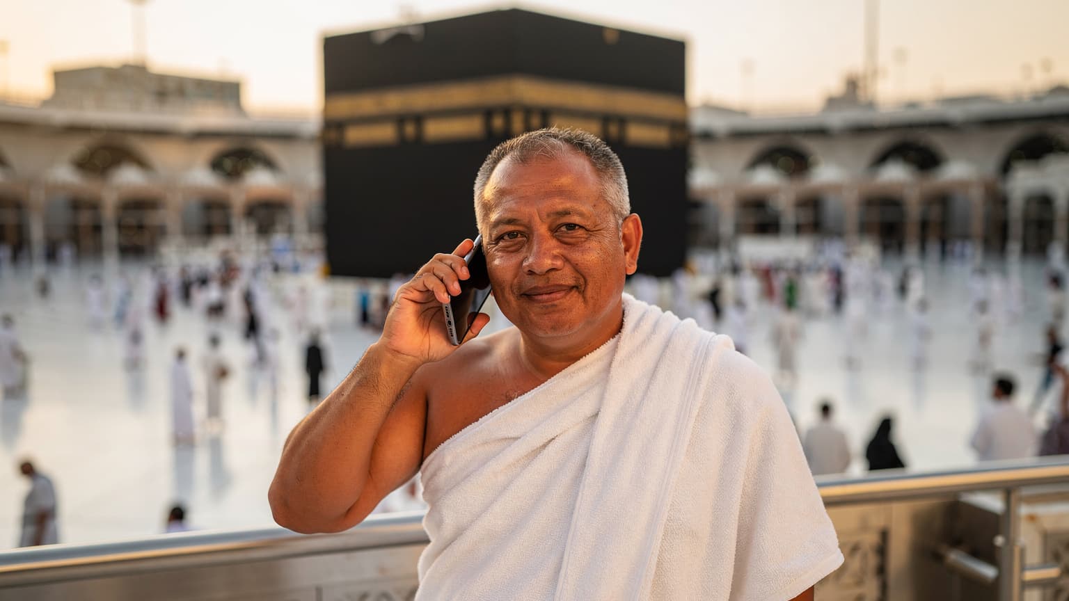 Seorang jemaah lelaki warga Malaysia sedang berbual melalui telefon pintar dengan latar belakang Masjidil Haram di Makkah pada waktu senja.