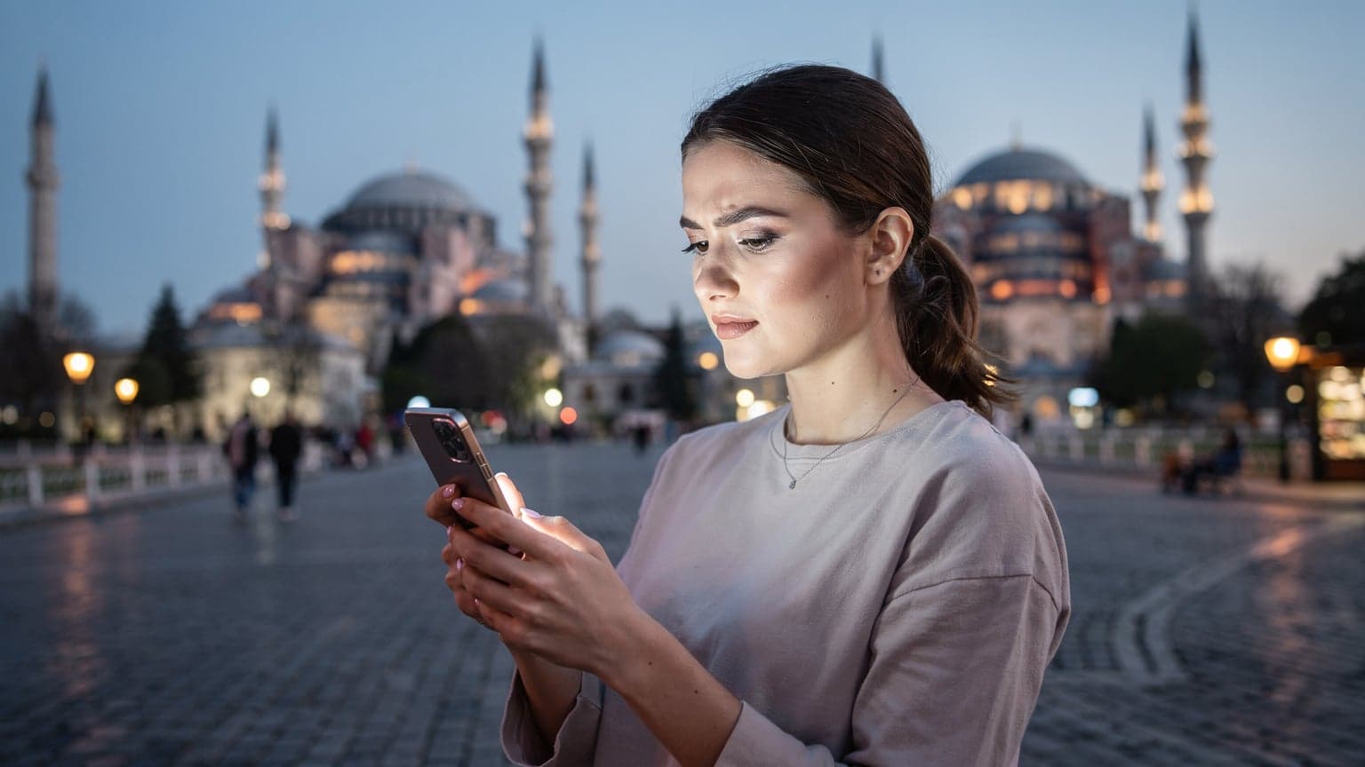 İstanbul'un Sultanahmet Meydanı'nda, mavi saatlerde, bir kadın telefonunda eSIM kurulumunu kontrol ediyor, arkasında Ayasofya ve Sultanahmet Camii'nin silüetleri beliriyor.