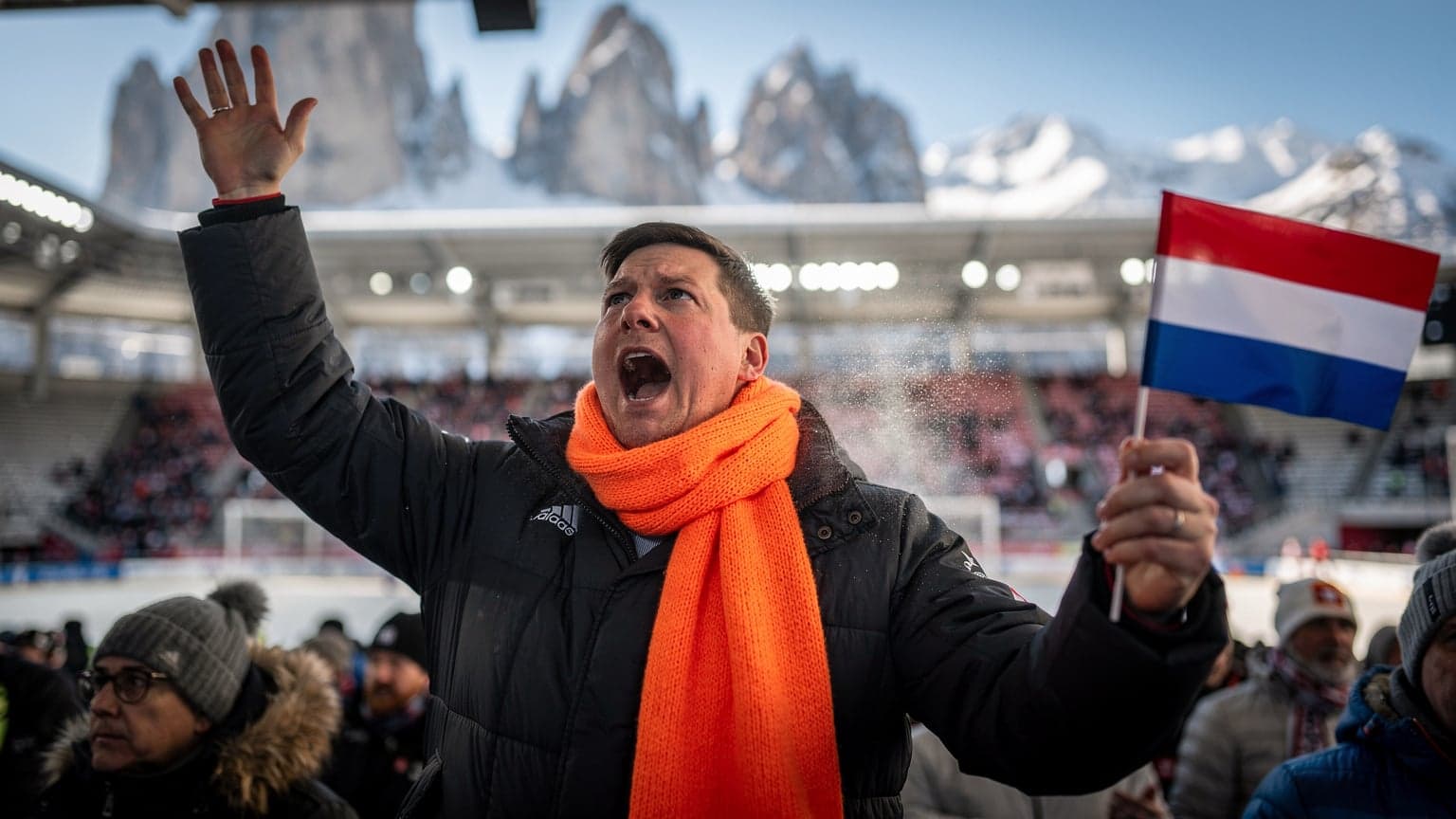 Een Nederlandse fan met oranje sjaal juicht enthousiast in het Olympisch Stadion van Cortina d'Ampezzo, met besneeuwde Dolomieten op de achtergrond.