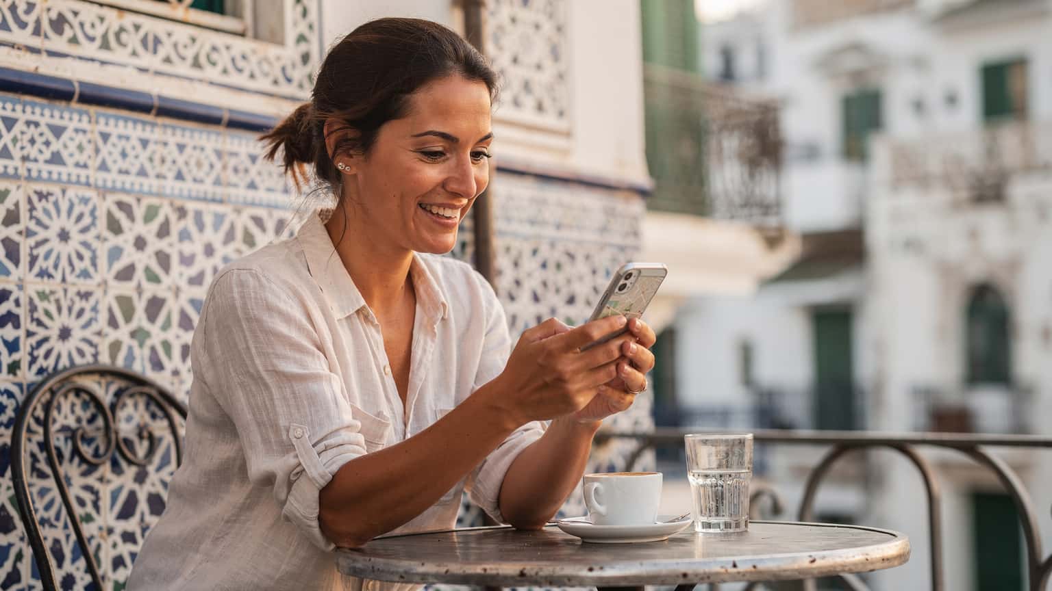 Une femme souriante tient son smartphone et consulte une carte en ligne, assise à la terrasse d'un café traditionnel dans la Casbah d'Alger, avec des bâtiments historiques en arrière-plan sous un ciel bleu clair.