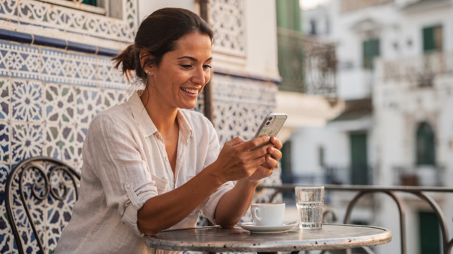 Une femme souriante tient son smartphone et consulte une carte en ligne, assise à la terrasse d'un café traditionnel dans la Casbah d'Alger, avec des bâtiments historiques en arrière-plan sous un ciel bleu clair.