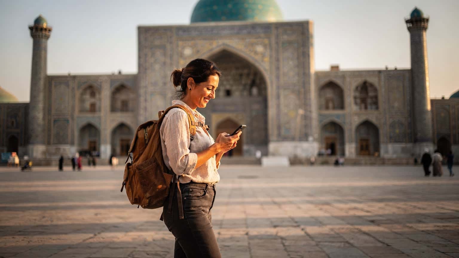 A seasoned female traveler, mid-thirties, smiling as she checks her phone for a map in front of the Registan in Samarkand, Uzbekistan, with intricate turquoise tilework visible.
