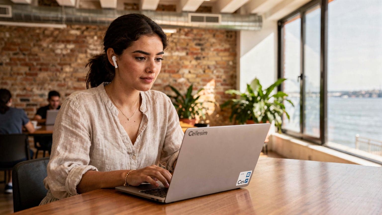 A digital nomad working on a laptop at a co-working space in Lisbon, with a focused expression and a Cellesim eSIM logo subtly visible on a sticker on her laptop.