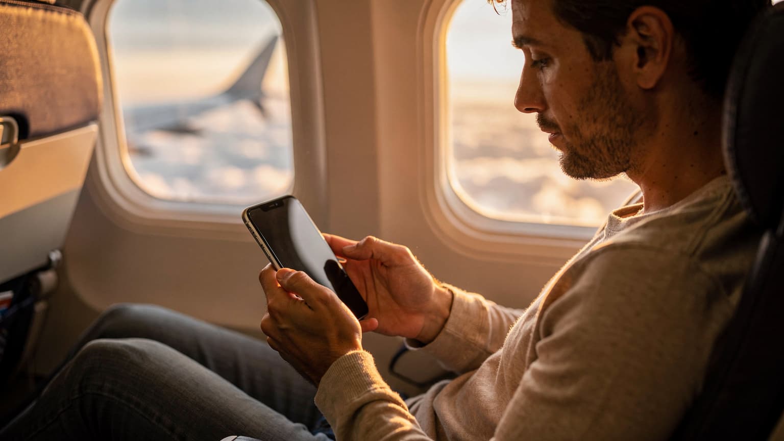 Eine Person betrachtet ihr iPhone 11 im Flugzeugsitz, während sie über den Wolken fliegt und die Morgensonne durch das Fenster scheint.