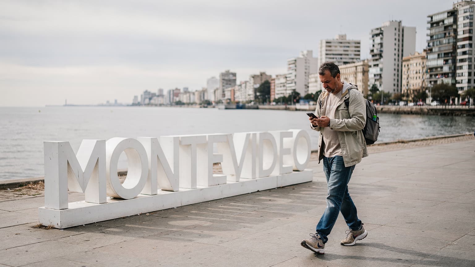 Un viajero revisa su teléfono mientras camina por la Rambla de Montevideo, con el icónico letrero de Montevideo al fondo y el Río de la Plata a su derecha, bajo un cielo parcialmente nublado.