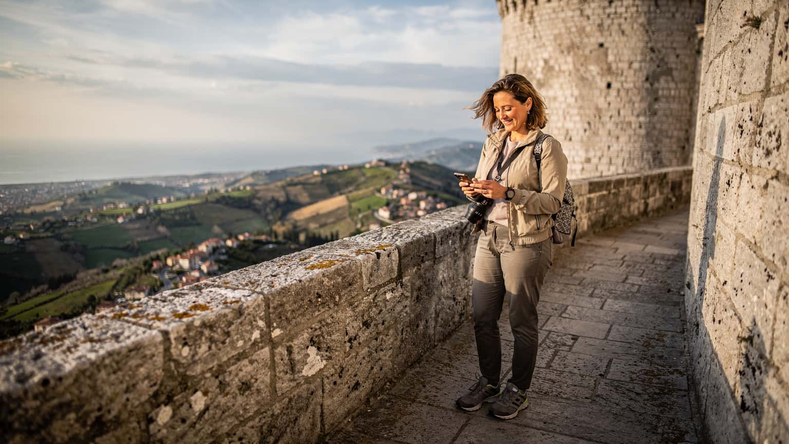 Una persona sta consultando il suo smartphone mentre si trova sulla Terza Torre, Montale, a San Marino, con una vista panoramica del paesaggio circostante al tramonto.