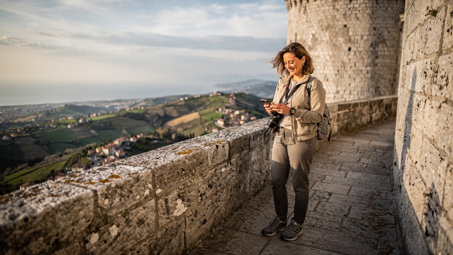 Una persona sta consultando il suo smartphone mentre si trova sulla Terza Torre, Montale, a San Marino, con una vista panoramica del paesaggio circostante al tramonto.