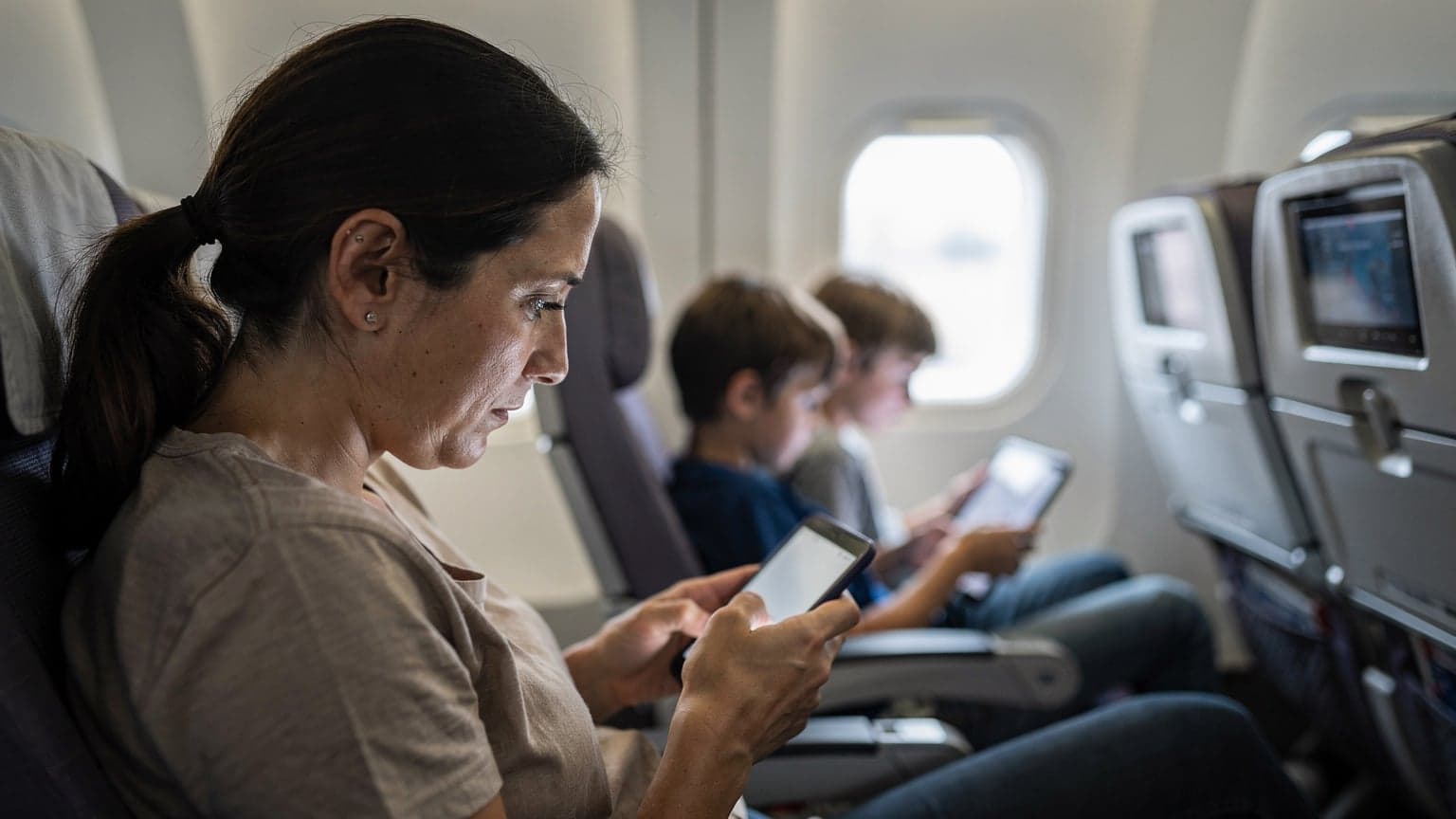 A parent looking at their smartphone with a slightly exasperated but determined expression, while two children in the background are engrossed in their tablets on a flight.