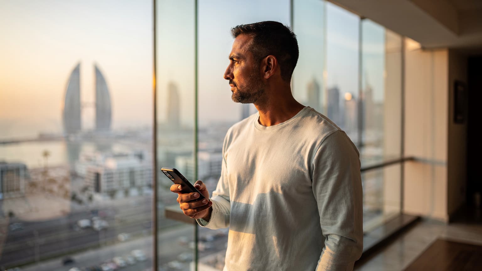 رجل يرتدي قميصًا خفيفًا يحمل هاتفًا ذكيًا ينظر إلى نافذة زجاجية كبيرة، تظهر من خلالها أبراج مدينة المنامة الحديثة عند الغروب، مع انعكاسات أضواء المدينة على الزجاج.