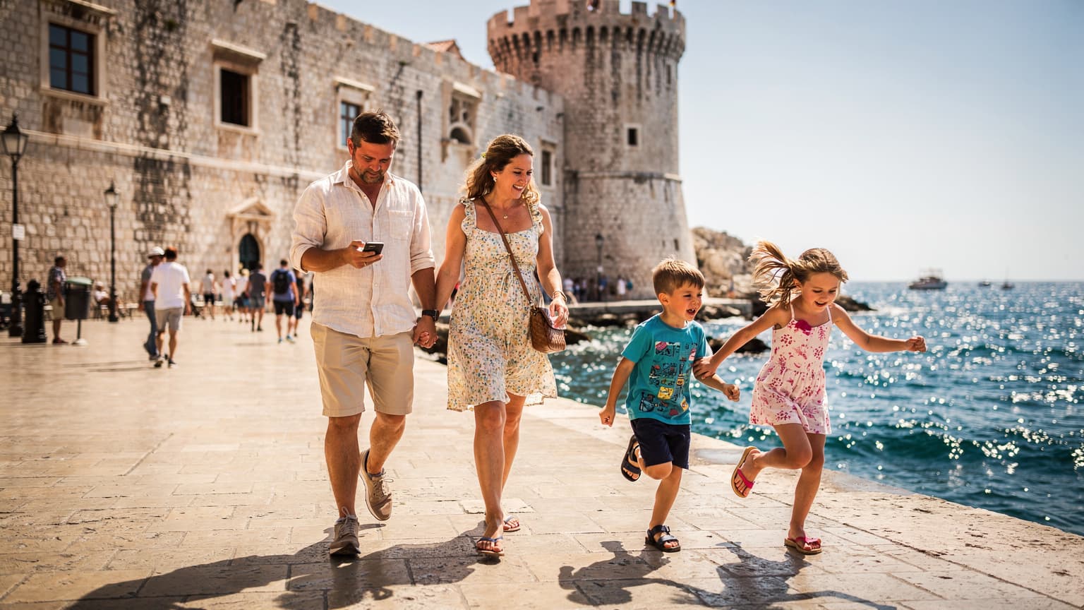 Eine Familie mit zwei Kindern, die lachend auf einer Promenade in Dubrovnik, Kroatien, spaziert, während der Vater auf sein Smartphone schaut.