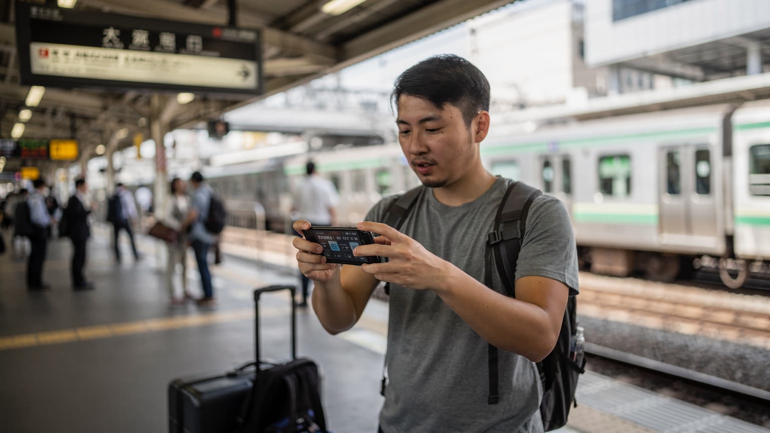 A person in a bustling Tokyo train station is looking at their smartphone screen, which displays a speed test application showing download and upload speeds, against a backdrop of commuters and a train pulling into the platform.
