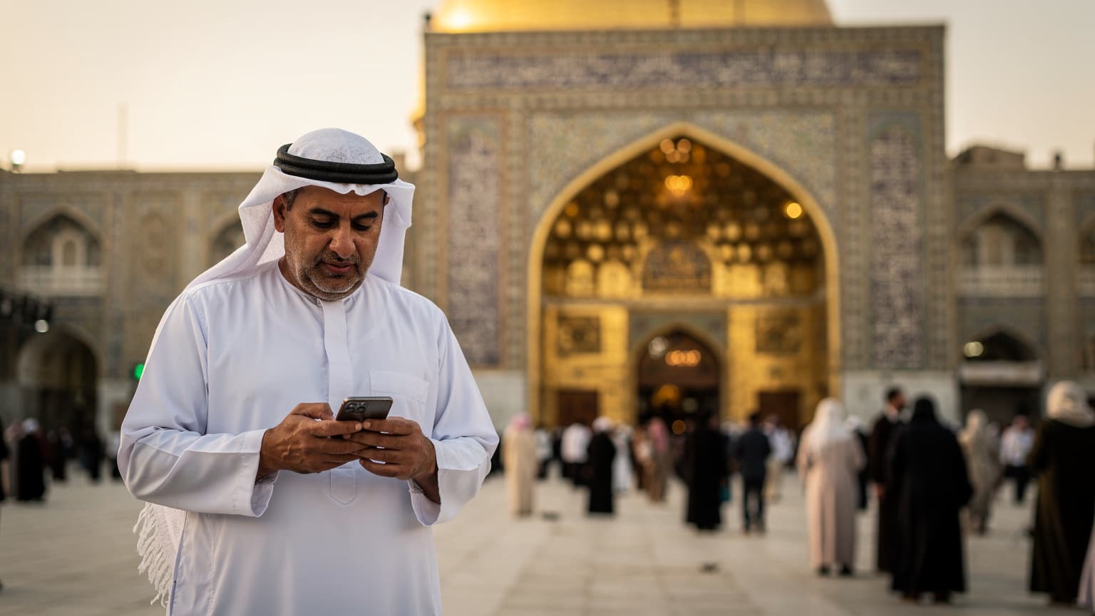 رجل عربي يرتدي الكوفية التقليدية يتصفح هاتفه الذكي أمام ضريح الإمام علي في النجف، العراق، مع إضاءة شمس الغروب الدافئة التي تبرز تفاصيل العمارة الإسلامية