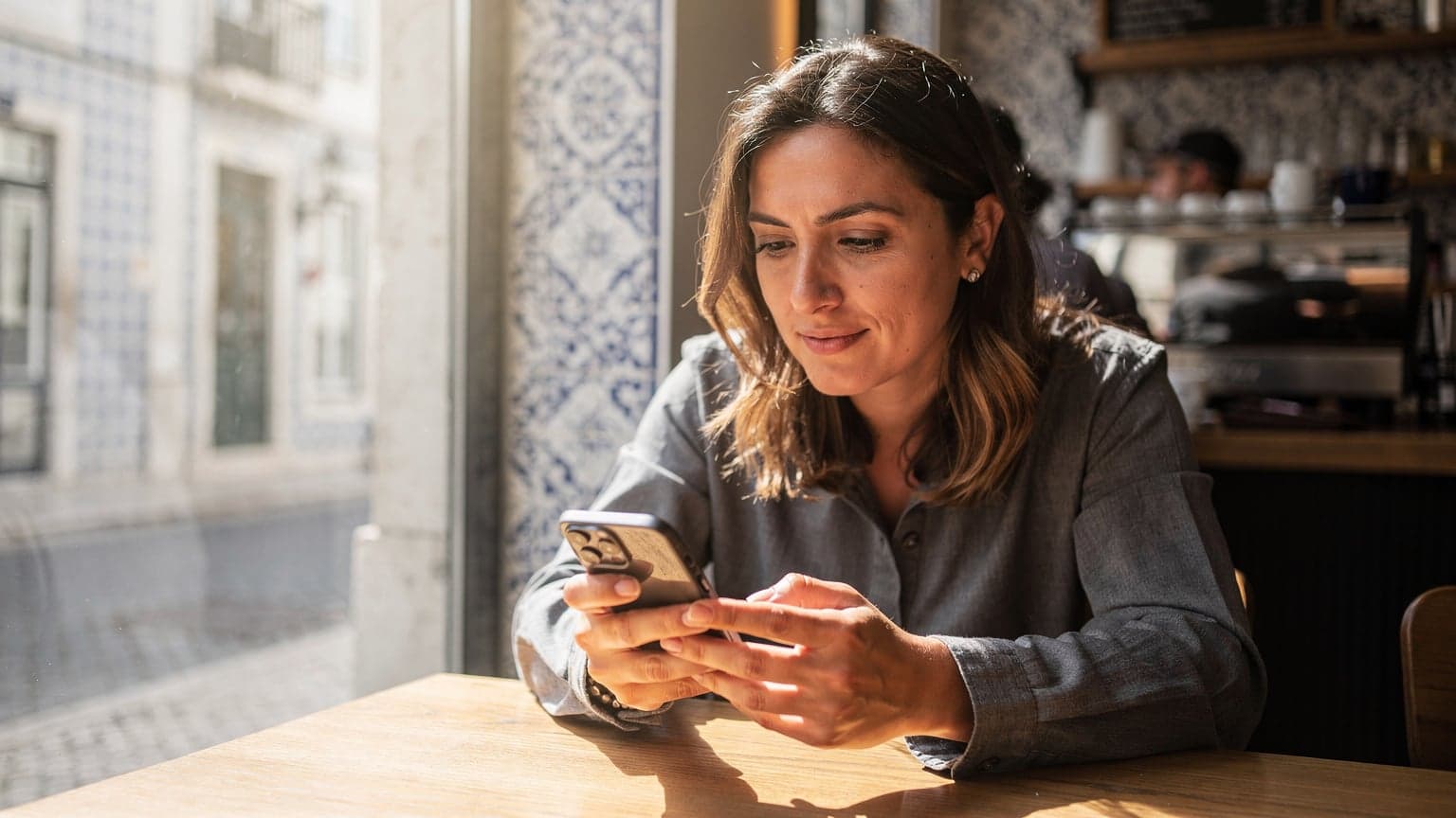 Una donna seduta al tavolo di un caffè di Lisbona, con il suo laptop aperto e uno smartphone in mano, mentre verifica la connessione eSIM per un viaggio imminente.