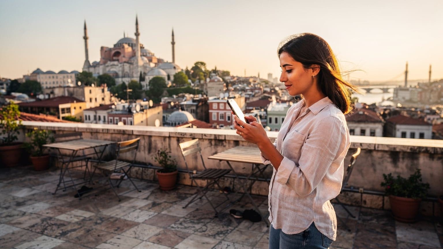 İstanbul'da tarihi bir binanın çatısında, elinde akıllı telefonuyla şehrin panoramik manzarasını izleyen genç bir kadın, telefonunda eSIM uygulamasını kontrol ediyor. Güneş batarken, sıcak turuncu ışıklar şehri aydınlatıyor.