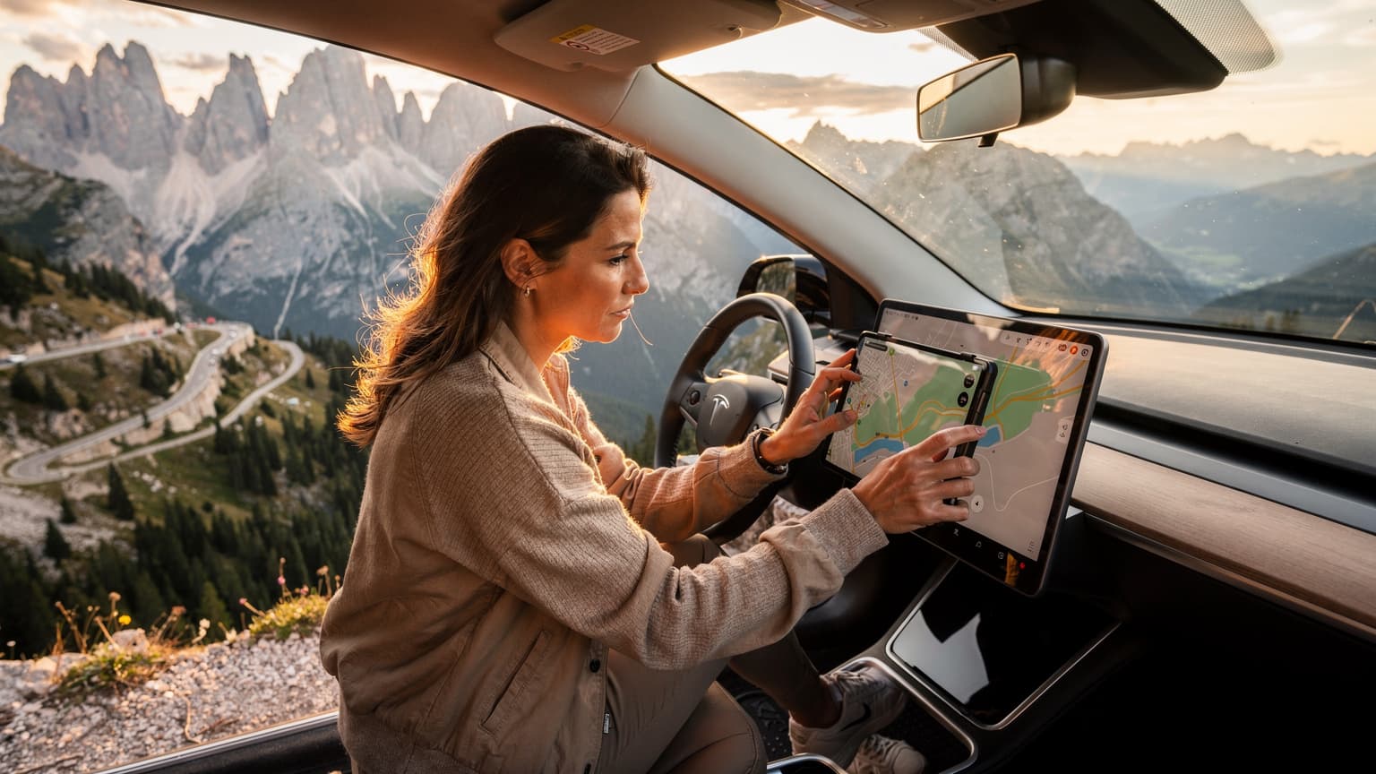 Eine Frau in einem modernen Elektroauto blickt auf ein Tablet, das eine Routenplanungs-App anzeigt, während sie in den Dolomiten parkt und die Berge im Hintergrund in der goldenen Abendsonne leuchten.