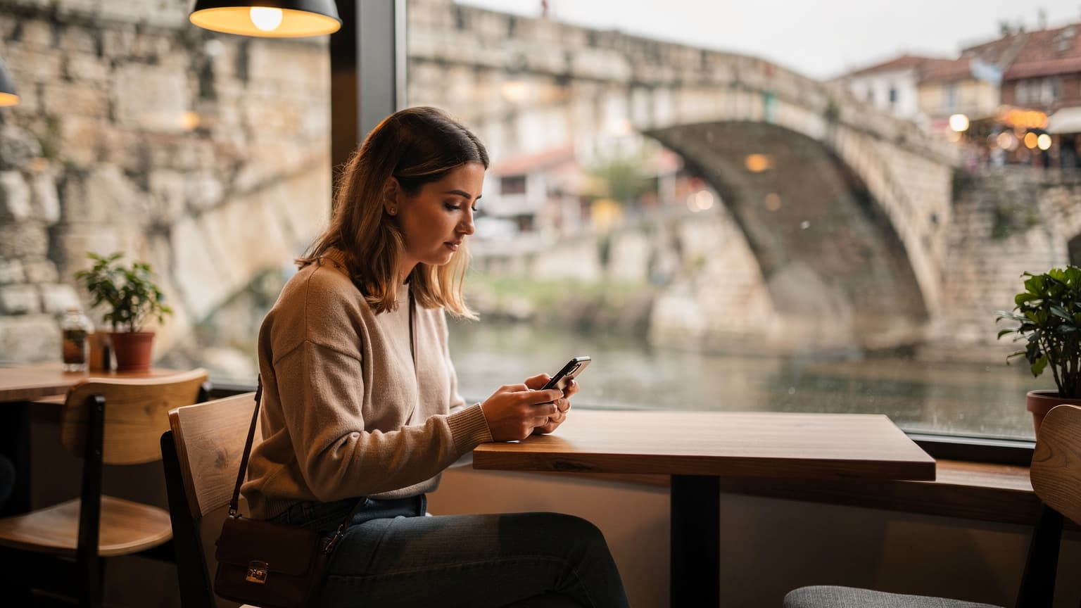 Eine Reisende sitzt in einem Café in Skopje, Nordmazedonien, und nutzt ihr Smartphone, während im Hintergrund die Steinbrücke zu sehen ist.