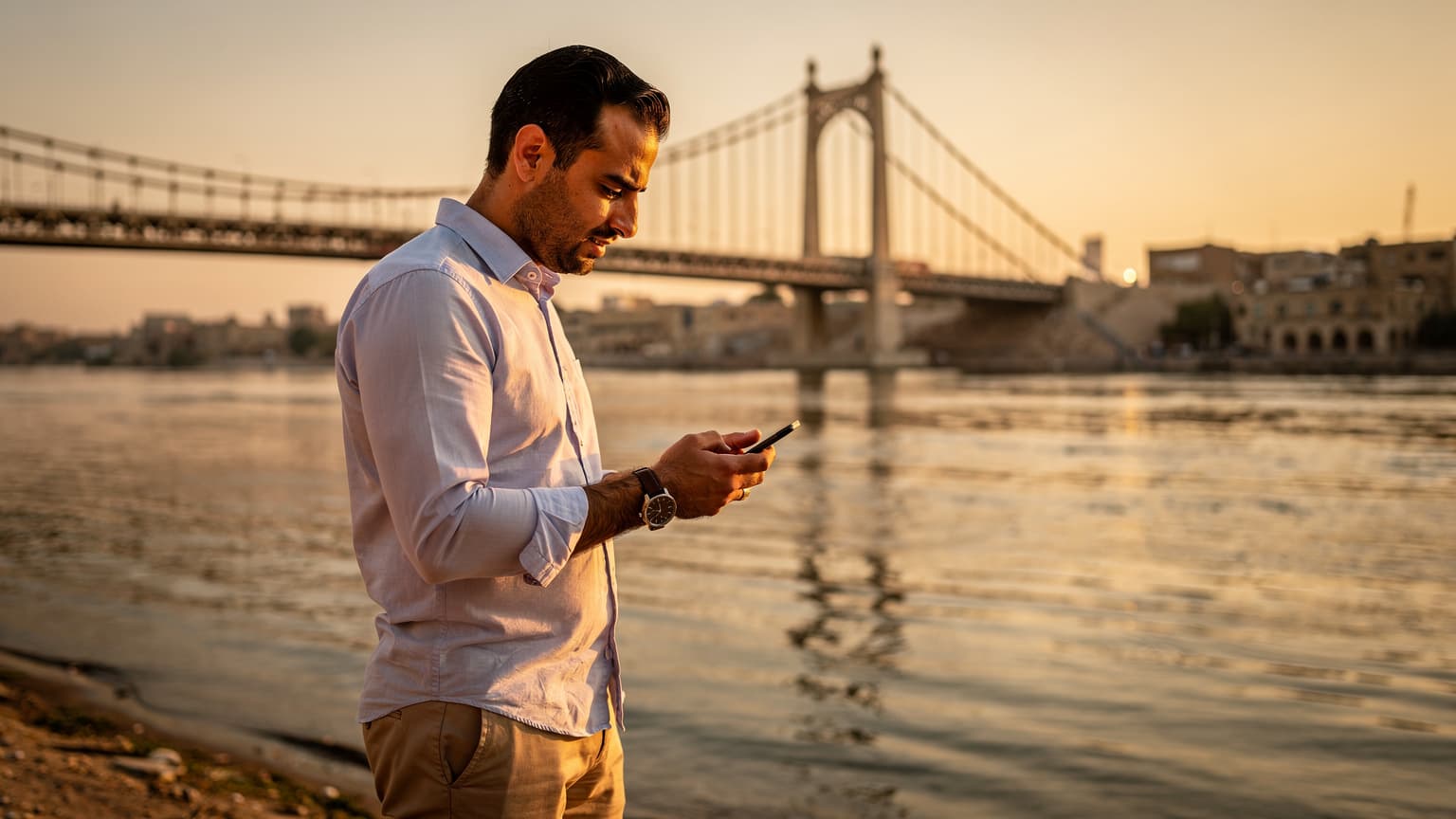 رجل يتفحص هاتفه الذكي على ضفة نهر دجلة بالقرب من الجسر المعلق في بغداد عند غروب الشمس.