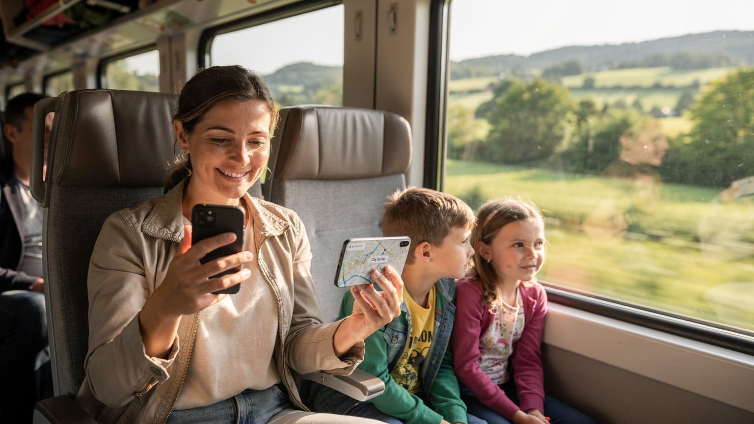 O mamă zâmbitoare, cu un telefon în mână, verificând o hartă pe ecran, în timp ce copiii ei, un băiat și o fată, privesc pe fereastra unui tren modern care traversează peisaje europene verzi. Lumina soarelui de dimineață pătrunde prin geam, creând o atmosferă caldă și primitoare.