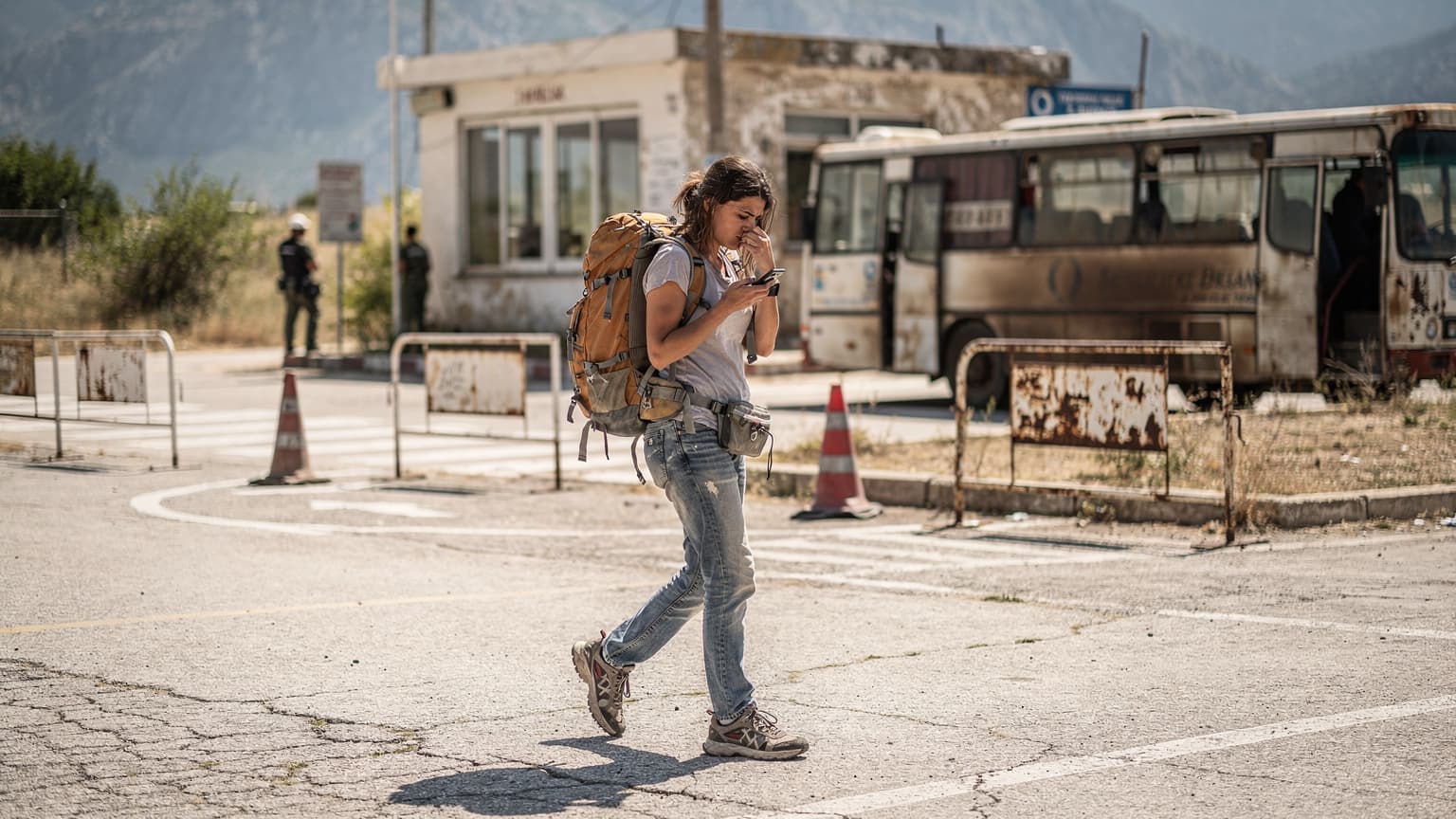 Eine junge Frau mit Rucksack überquert zu Fuß einen Grenzübergang auf dem Balkan, im Hintergrund eine verwitterte Zollstation und ein alter Bus, während sie auf ihr Smartphone blickt, um die eSIM-Aktivierung zu überprüfen.