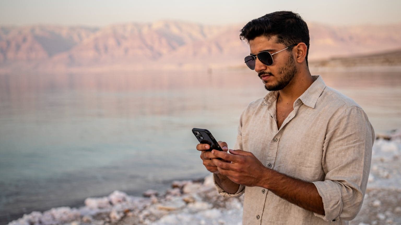 شاب عربي يرتدي نظارة شمسية ويحمل هاتفاً ذكياً في يده، يتصفح خريطة على شاطئ البحر الميت مع الجبال الوردية في الخلفية.