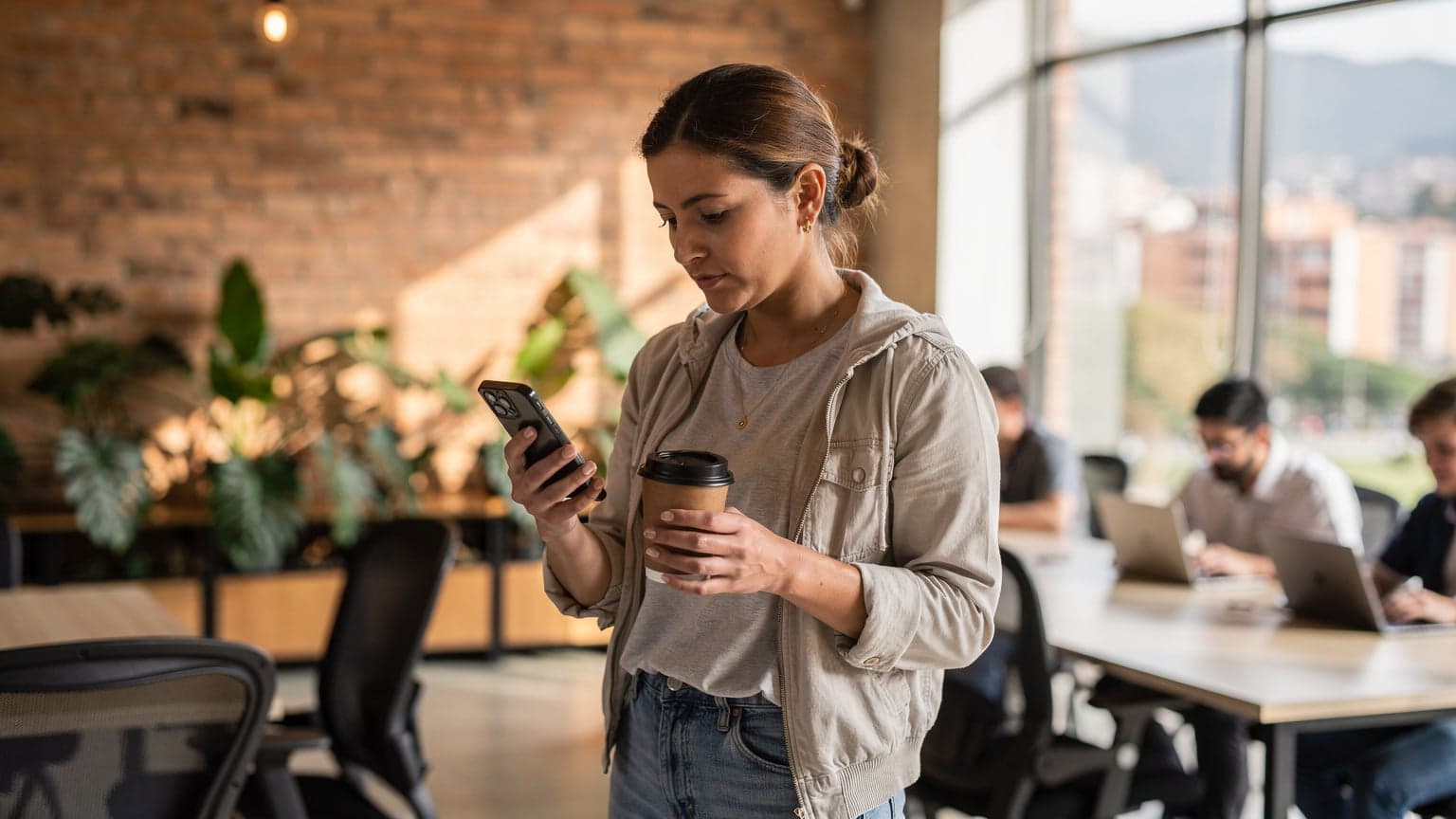 A digital nomad sits at a wooden table in a busy co-working space in Medellín, Colombia, looking at her smartphone while sipping coffee, with her laptop open next to her.