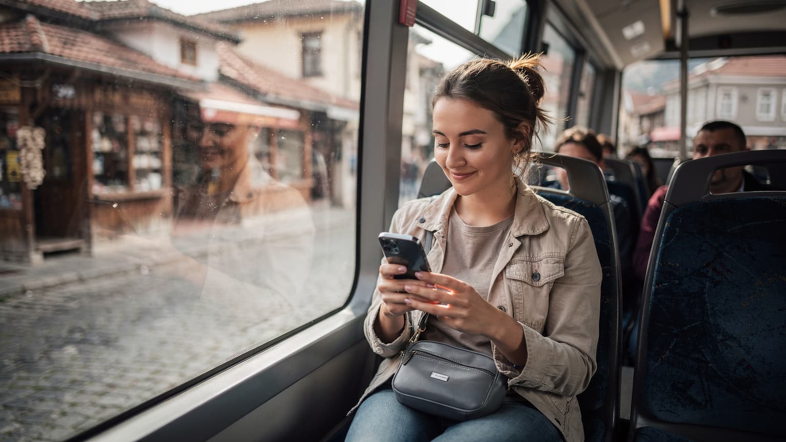 En kvinna sitter på en buss i Sarajevo, tittar på sin telefon med ett leende, med en ryggsäck bredvid sig.