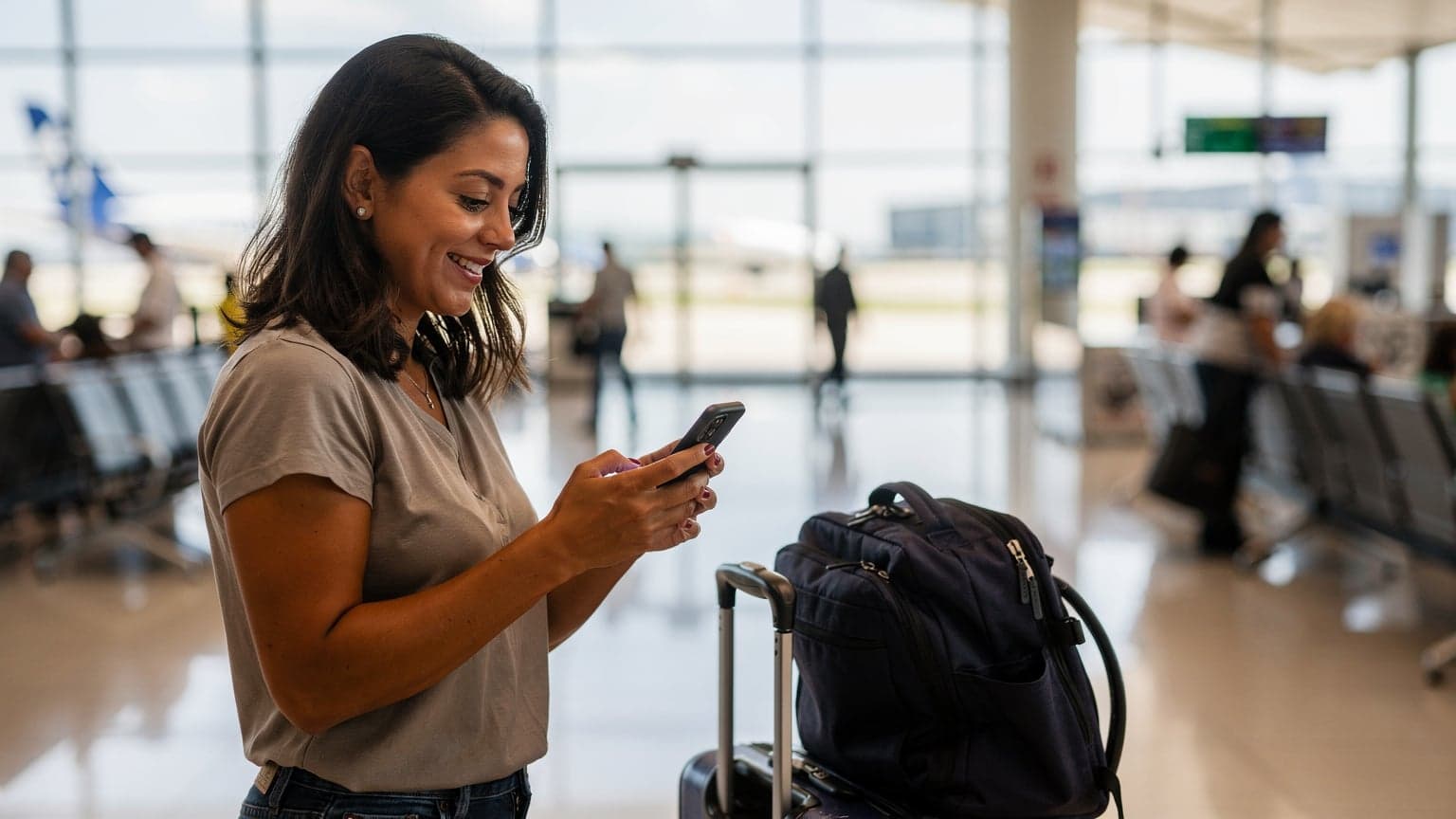 Uma mulher sorridente em um aeroporto movimentado, usando seu smartphone para verificar informações de voo, com malas de viagem ao seu lado.