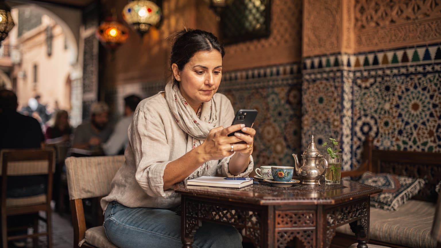 Une voyageuse française utilise Google Traduction sur son smartphone, assise à une table en bois dans un café traditionnel animé de la vieille ville de Marrakech, avec des motifs géométriques et des théières en laiton en arrière-plan.