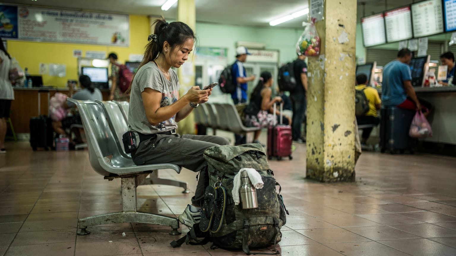 A budget backpacker checks their phone at a bustling bus station in Chiang Mai, Thailand, with a large backpack at their feet.