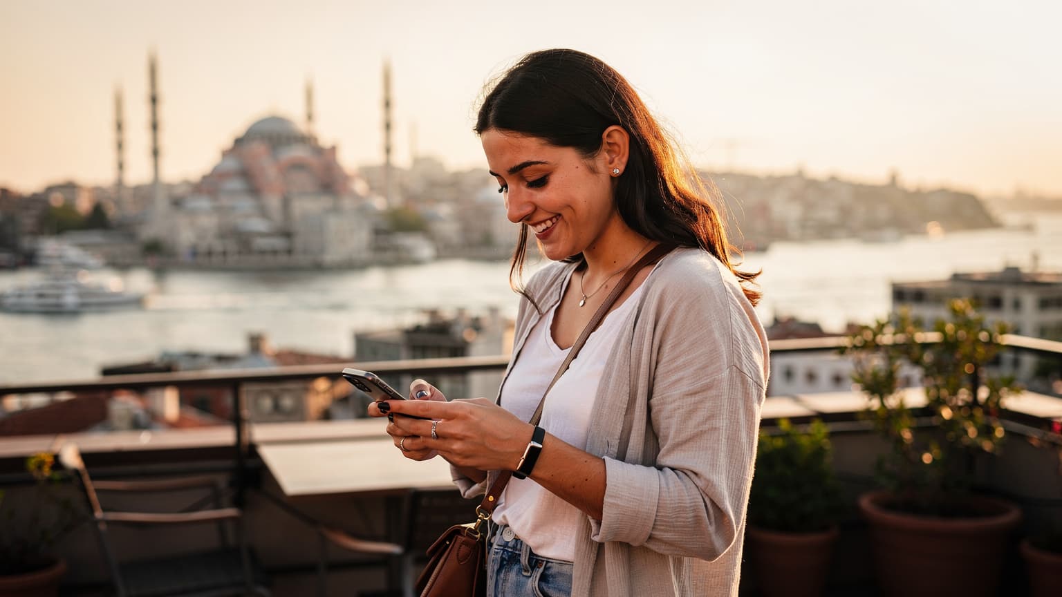 O tânără călătoare zâmbește în timp ce privește ecranul telefonului său, având în fundal o panoramă a Bosforului cu Aya Sofia și Moscheea Albastră la apus, în Istanbul.