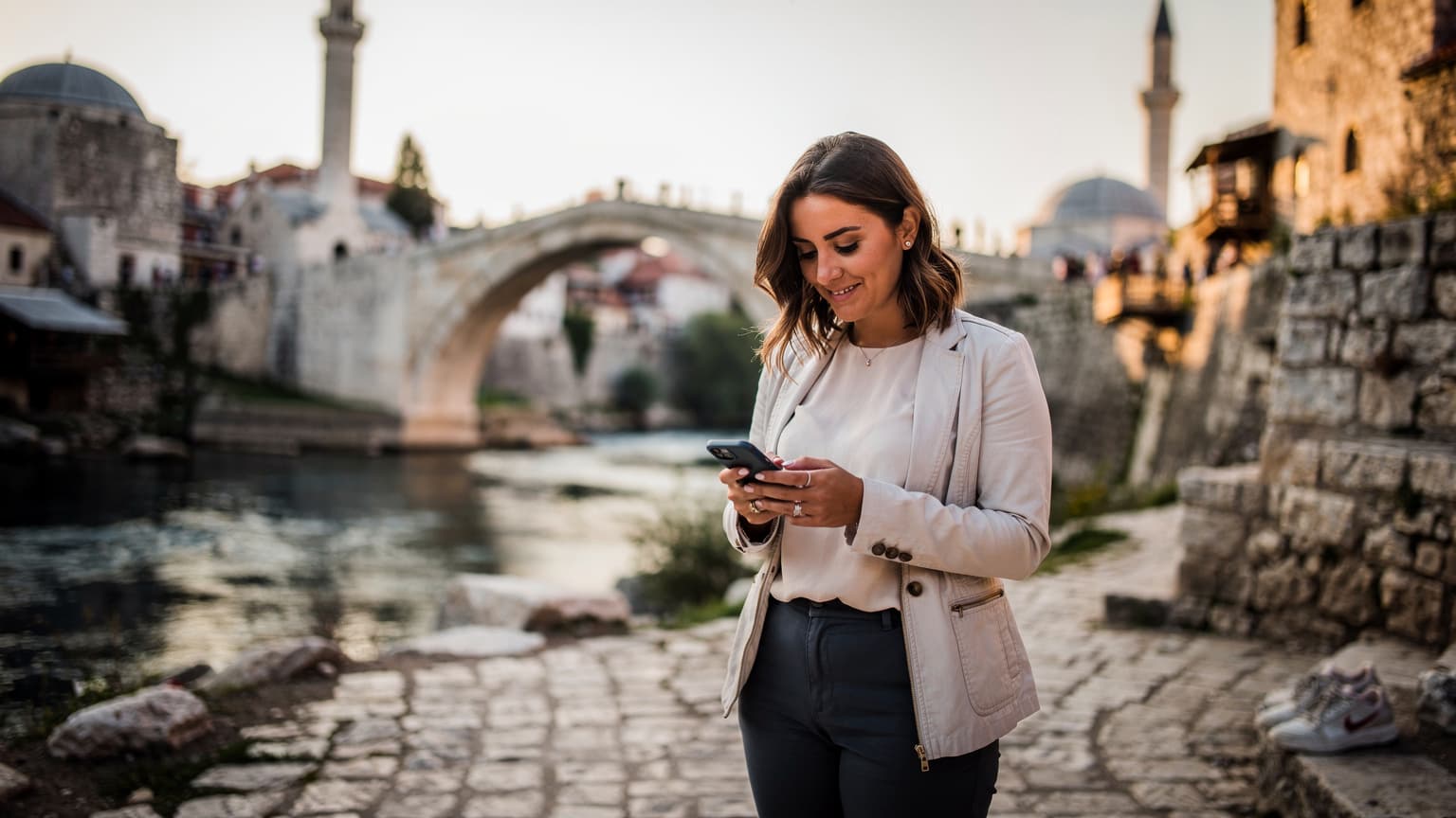 En kvinna tittar på sin smartphone med en moské i Mostar i bakgrunden, med ett lätt leende på läpparna.