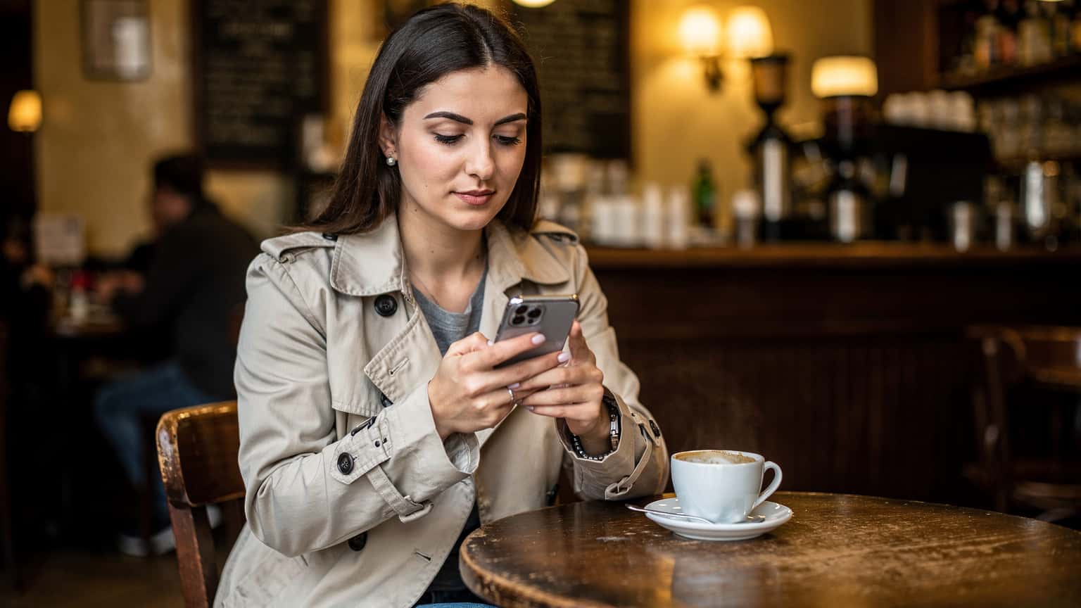Une voyageuse assise dans un café branché du quartier de la Croix-Rousse à Lyon, buvant un café tout en consultant son téléphone mobile pour planifier son itinéraire de voyage, son sac à dos posé à côté d'elle.