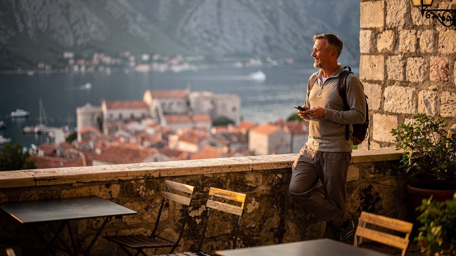 Een reiziger bekijkt de kustlijn van Kotor, Montenegro, vanaf een terrasje met een mobiele telefoon in de hand, terwijl de zon langzaam ondergaat.