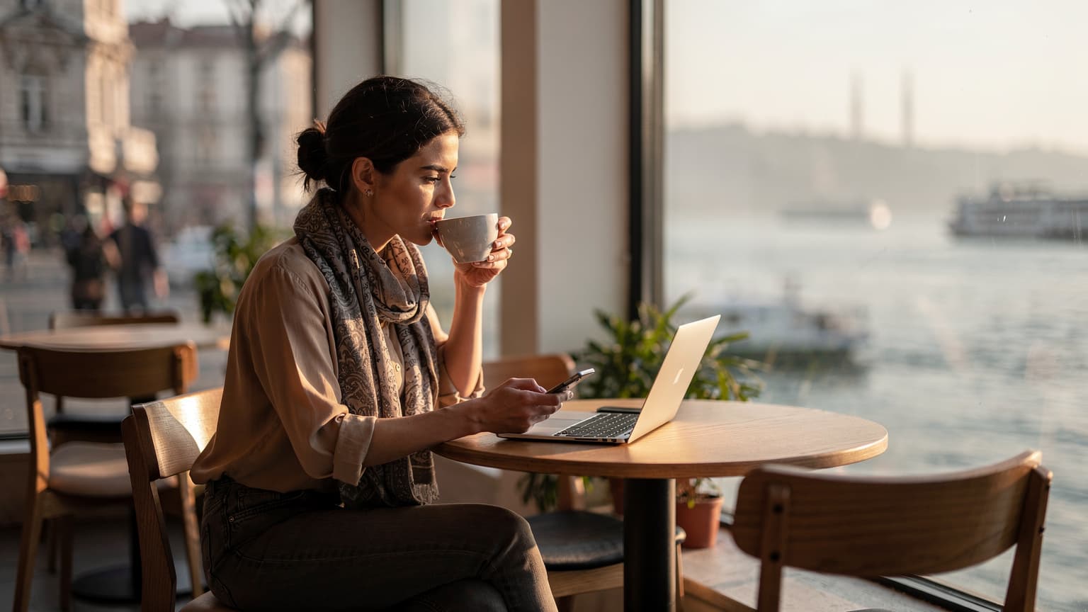 Bir genç kadın, İstanbul Boğazı'na bakan modern bir kafede laptopu ve akıllı telefonuyla çalışırken kahve yudumluyor.