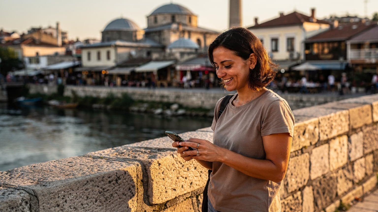 Kobieta korzystająca ze smartfona z widokiem na panoramę Skopje i rzekę Wardar, z charakterystycznymi mostami i architekturą, w tle.