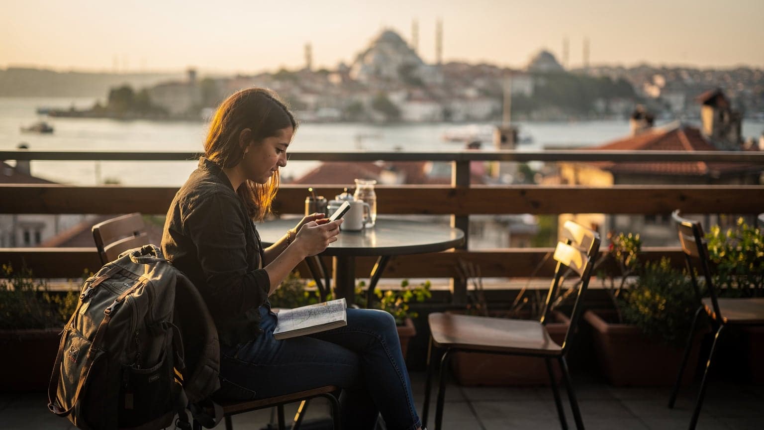O femeie tânără cu rucsac, verificându-și telefonul la o masă exterioară, cu vedere la Bosfor, în Istanbul, la apus.