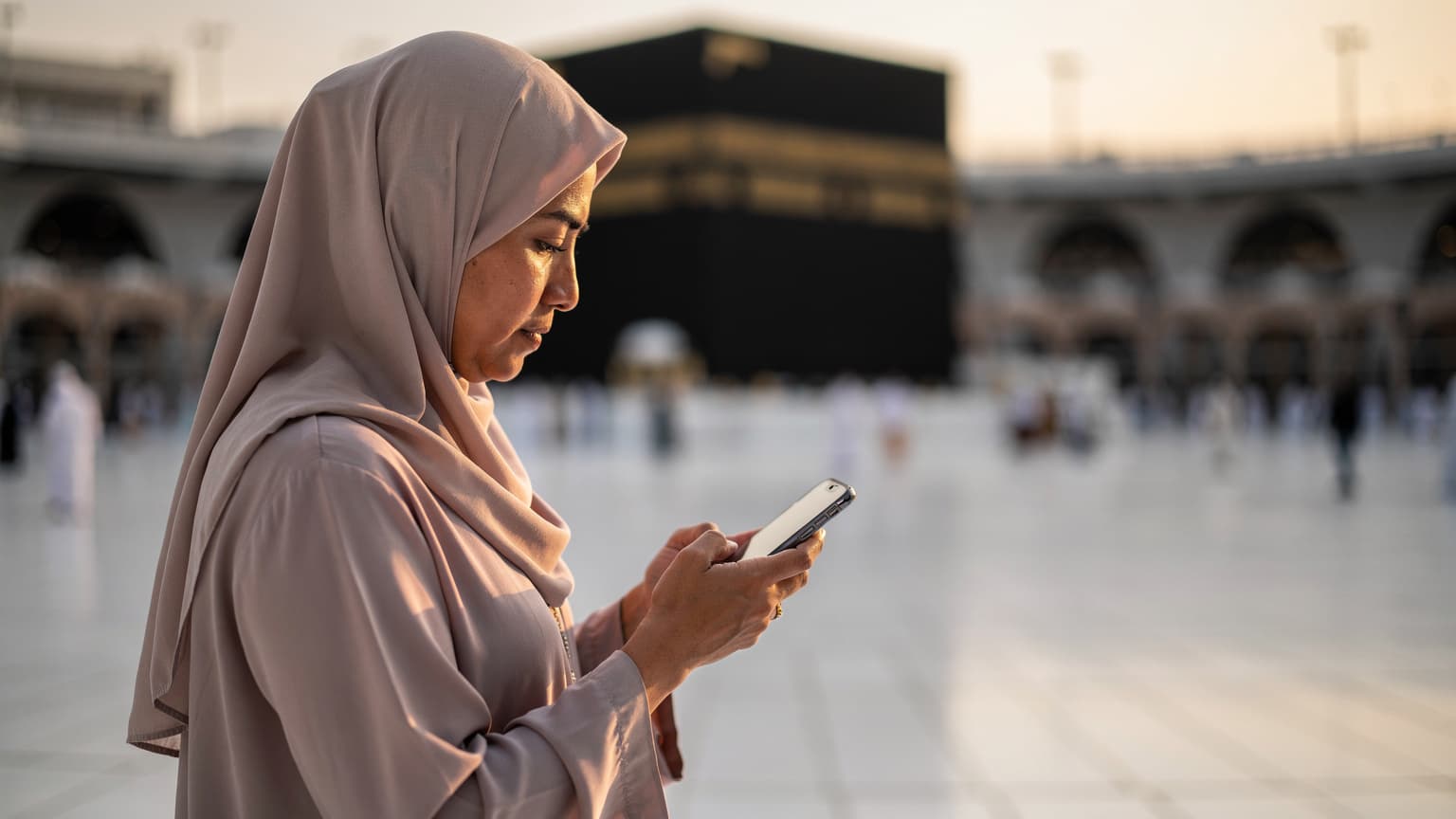 Seorang wanita Muslimah sedang melihat telefonnya di Masjidil Haram, Mekah, dengan Kaabah kelihatan samar di latar belakang, pada waktu subuh.