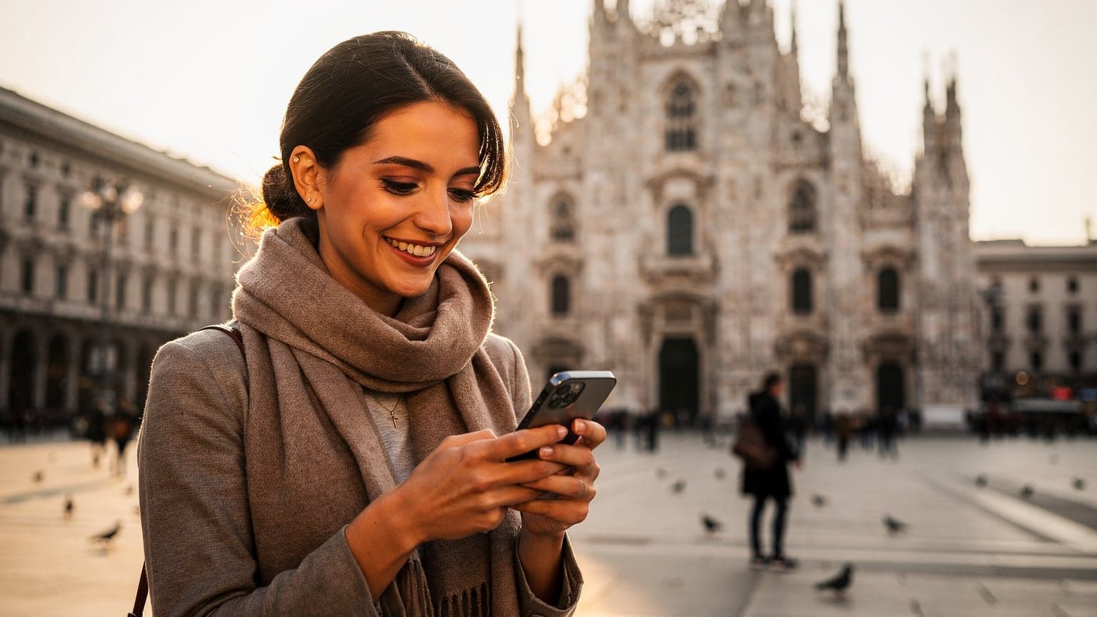 Una viaggiatrice sorride mentre naviga sul suo smartphone, con il Duomo di Milano in lontananza e il sole che illumina la piazza.