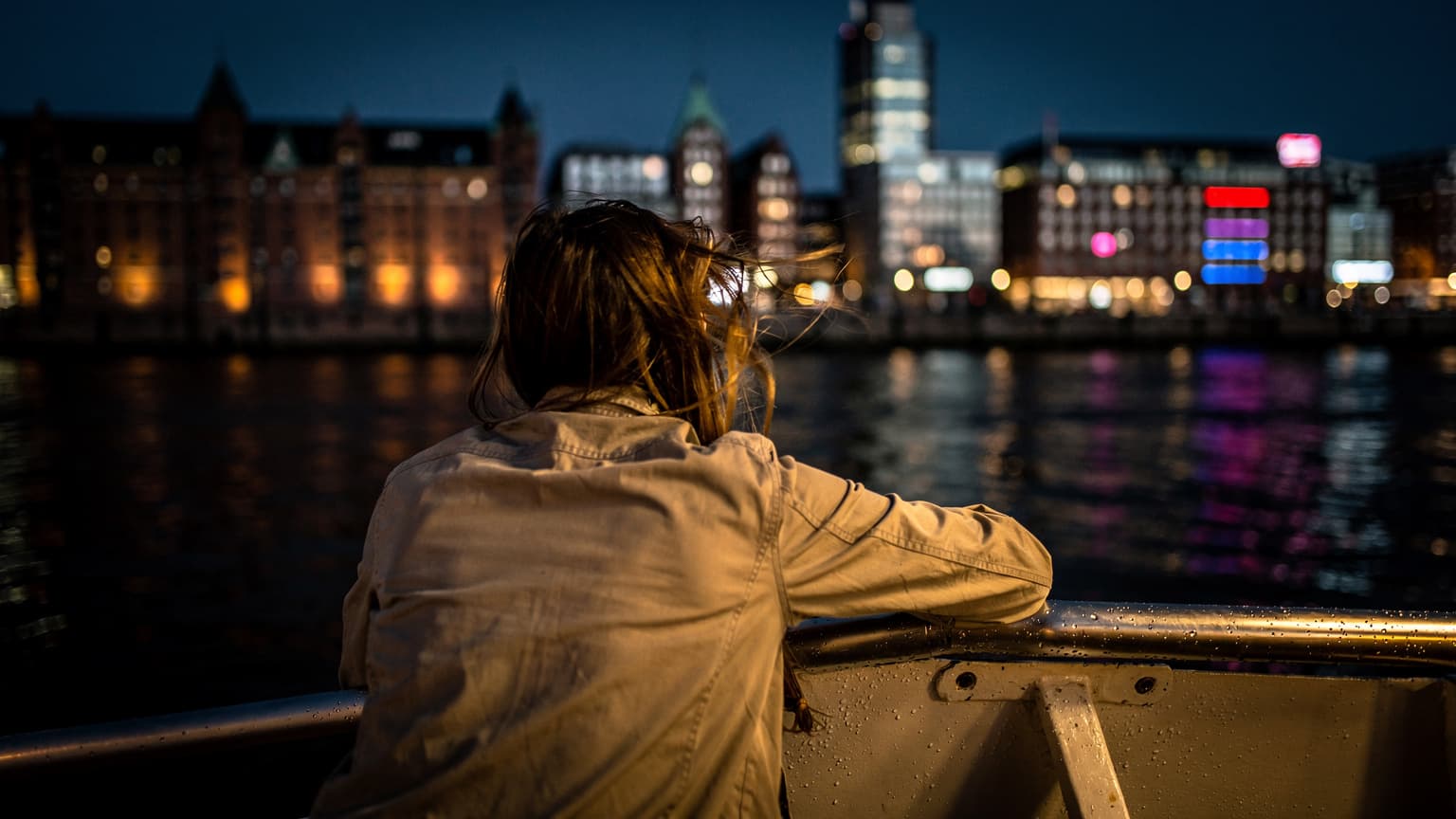 Eine Person lehnt nachts auf dem Deck einer Fähre im Hamburger Hafen, der Wind weht durch ihr Haar, im Hintergrund leuchten die Lichter des Fischmarkts und der Speicherstadt.