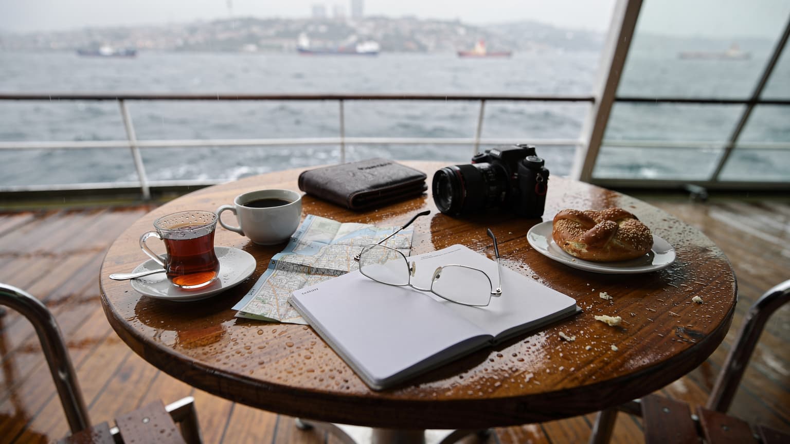 Una veduta dall'alto di un tavolo di un caffè sul ponte di un traghetto che attraversa il Bosforo in una giornata piovosa e grigia, con una tazza di caffè turco, un simit e una mappa cartacea delle isole Princes, con un telefono posato a faccia in giù.
