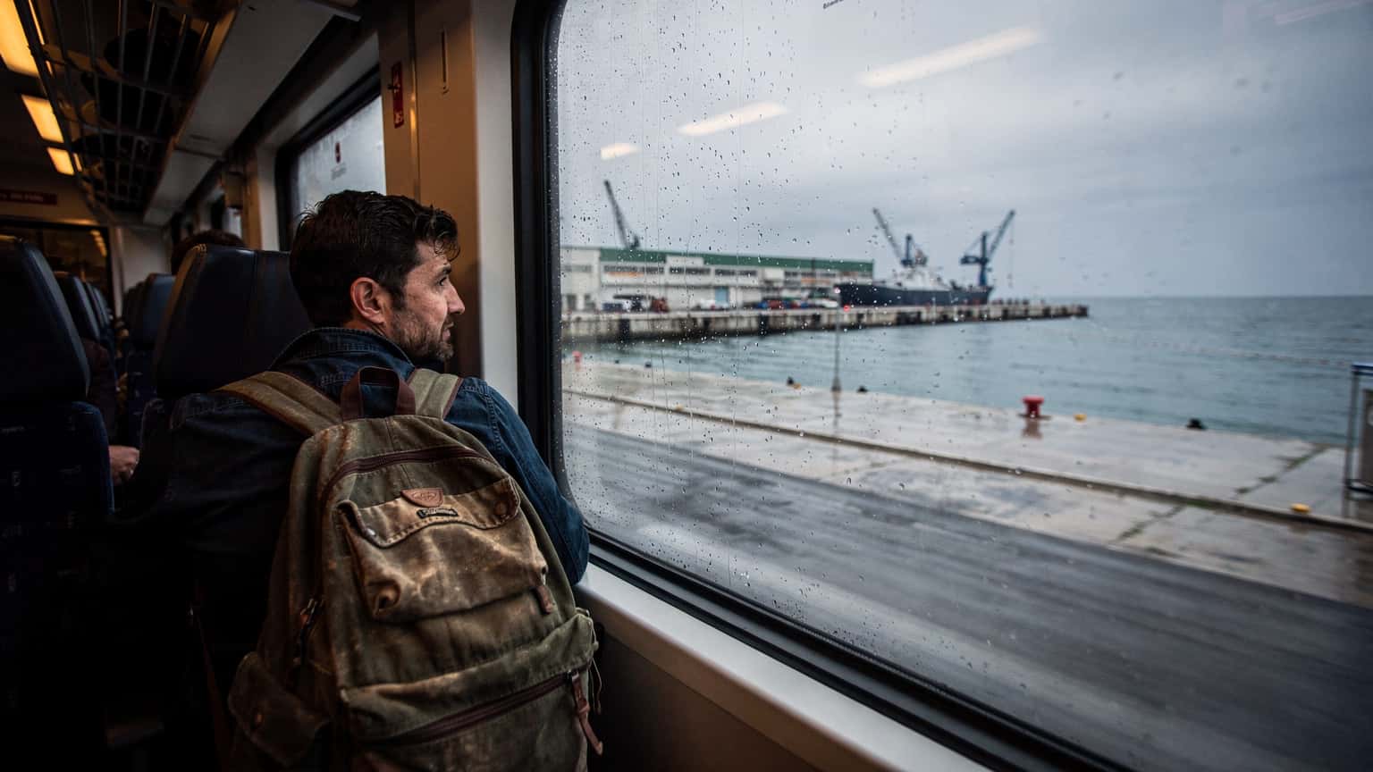 Rejsende med rygsæk ser ud ad vinduet på et tog, der kører gennem regnvejr langs havnefronten i Durrës, Albanien. Gråt lys og våde overflader, dokumentarisk stil.