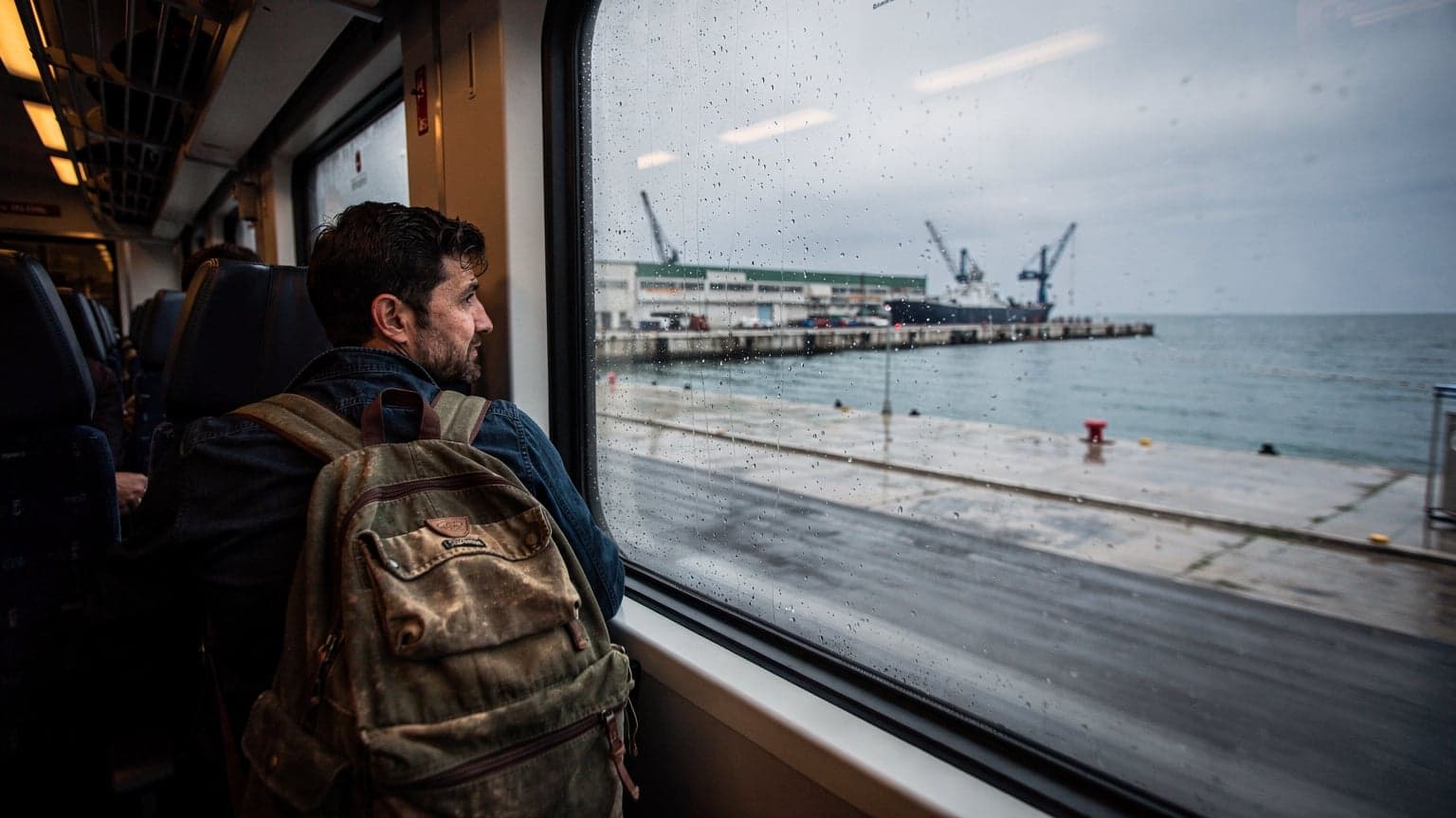 Rejsende med rygsæk ser ud ad vinduet på et tog, der kører gennem regnvejr langs havnefronten i Durrës, Albanien. Gråt lys og våde overflader, dokumentarisk stil.