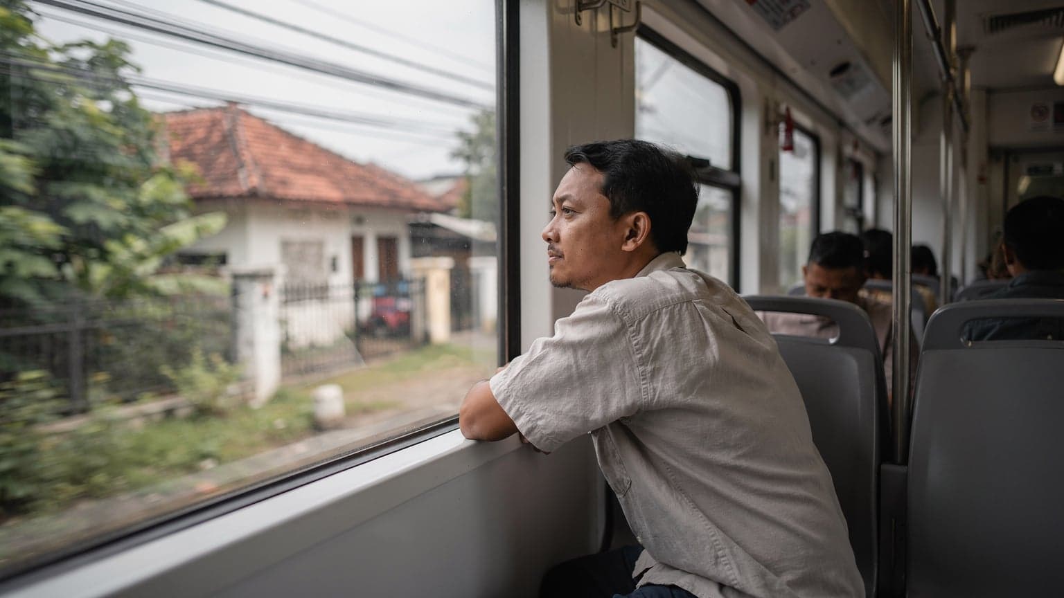 A traveler gazes out of a train window, watching the suburban landscape of Jakarta pass by under a soft, overcast morning sky, their backpack resting on the seat beside them.
