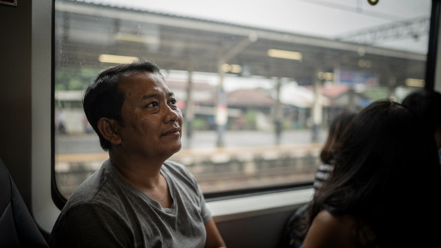 A passenger looking out of a train window in Jakarta during a soft overcast mid-morning, with their backpack on the seat next to them.