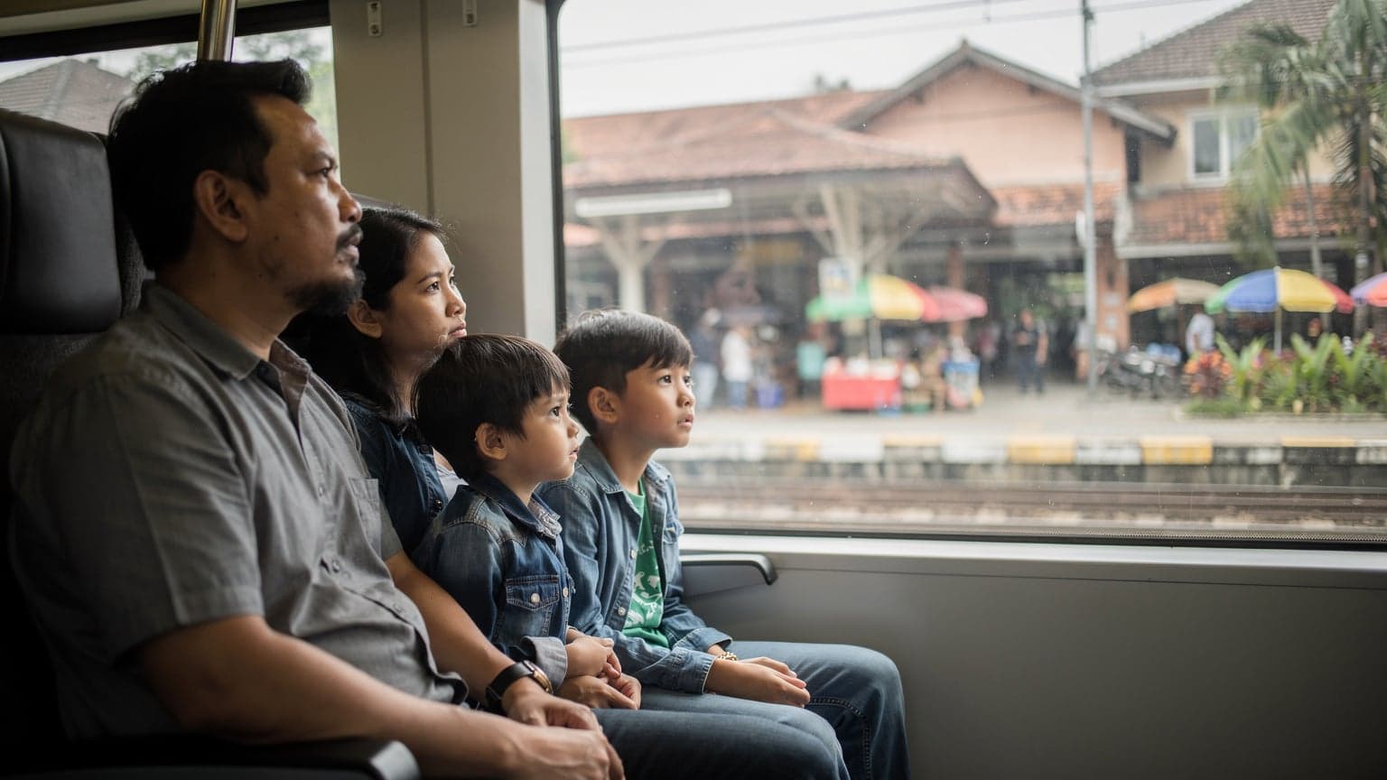 A family of four, two children aged 8 and 11, and their parents, gazing out of a train window in Jakarta, Indonesia, as a suburban rail platform with a distinctive local architectural style slides past in the mid-morning light. Their carry-on bags are on the seats beside them.